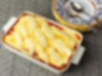 Baked lasagna in a white dish, topped with melted cheese. It sits on a patterned surface beside colorful plates, a fork, and a spoon.