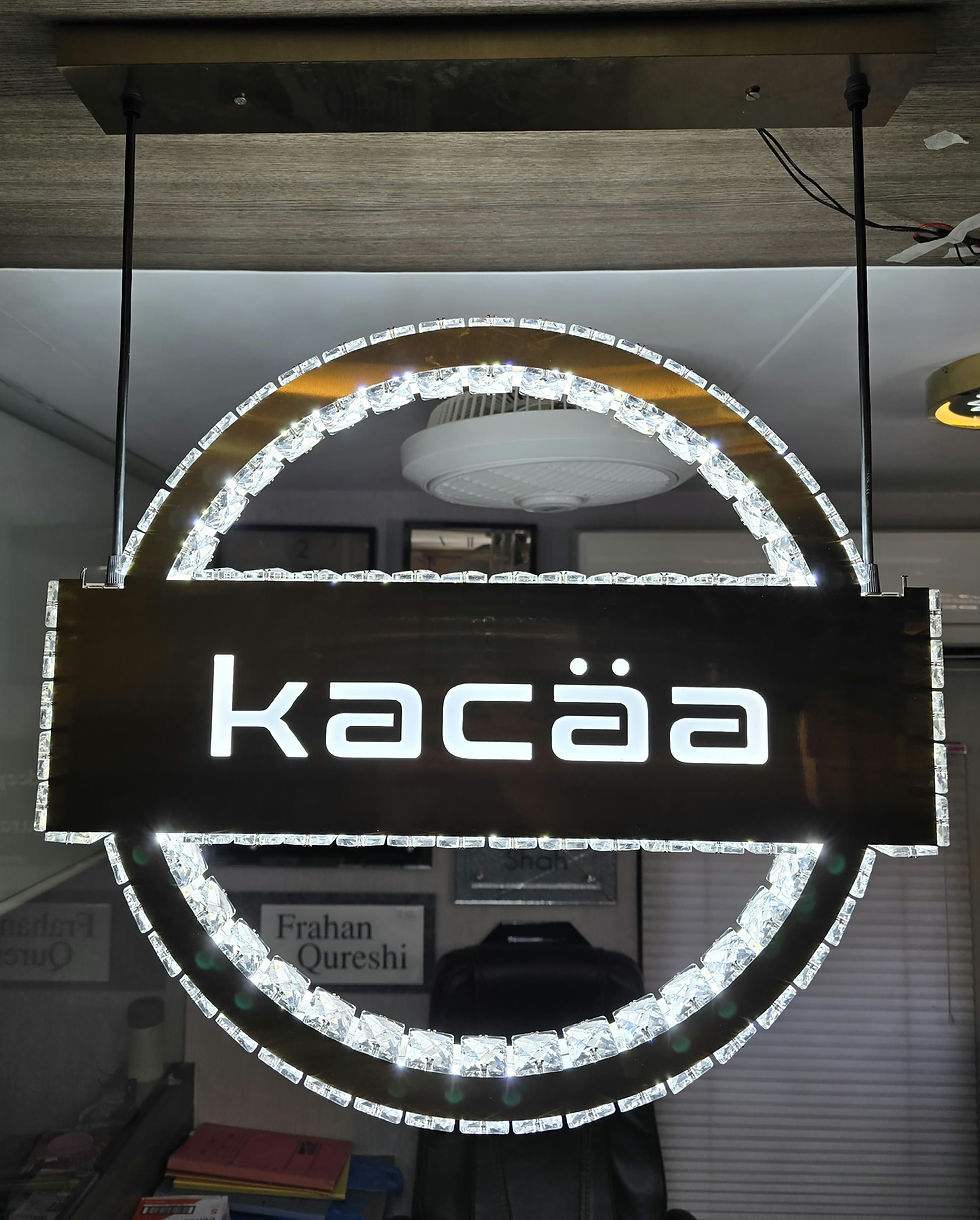 A circular, suspended name plate with a bronze or brass metallic finish. The circular frame is embedded with numerous clear, faceted crystal-like pieces that are brightly illuminated, creating a halo effect. The center features a black background with the white, illuminated logo "kacaa." This striking, illuminated name plate design is suspended from above.