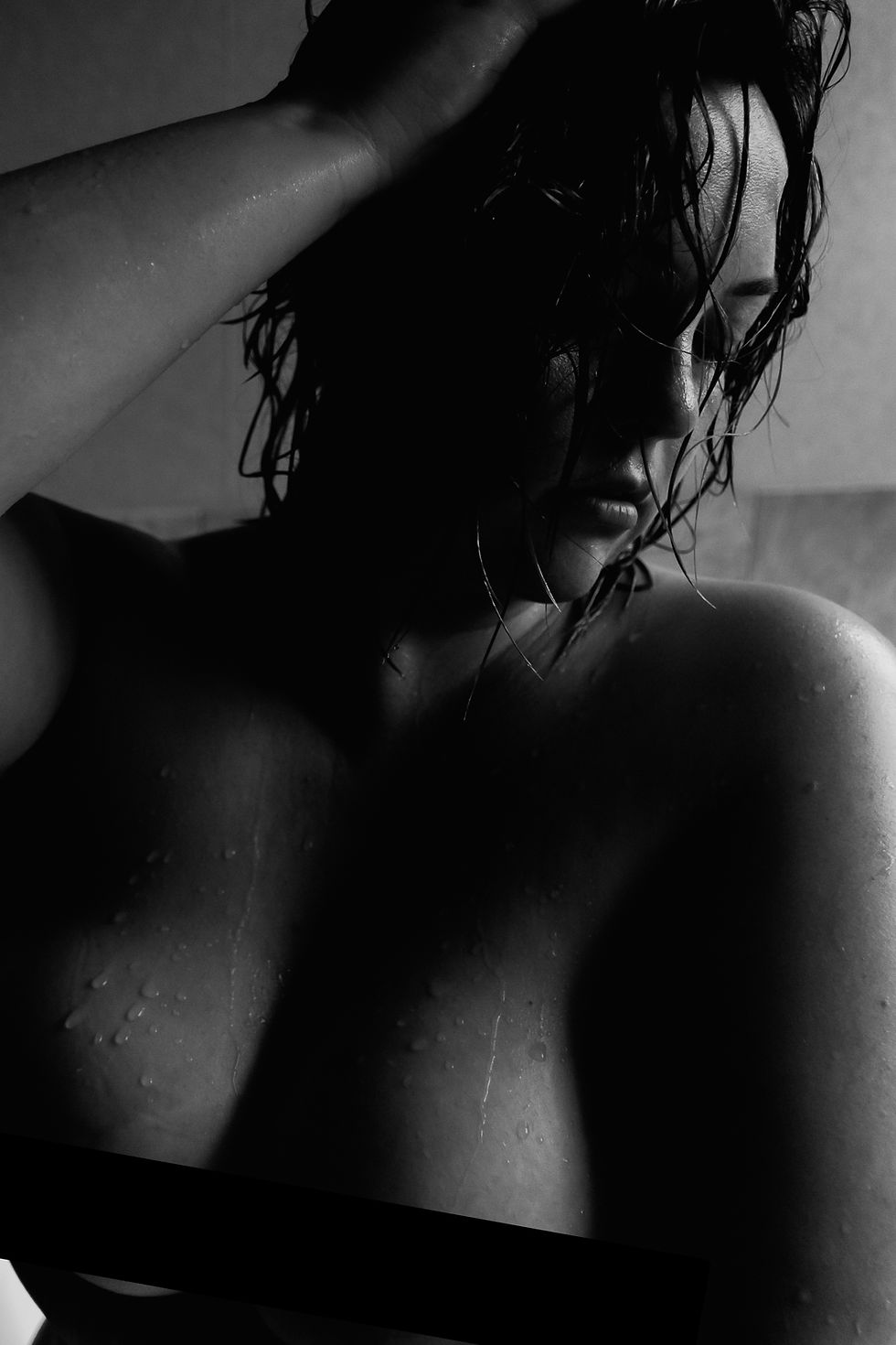 A woman with wet hair poses in dim light, hand on head, water droplets on skin, creating a dramatic and contemplative mood.