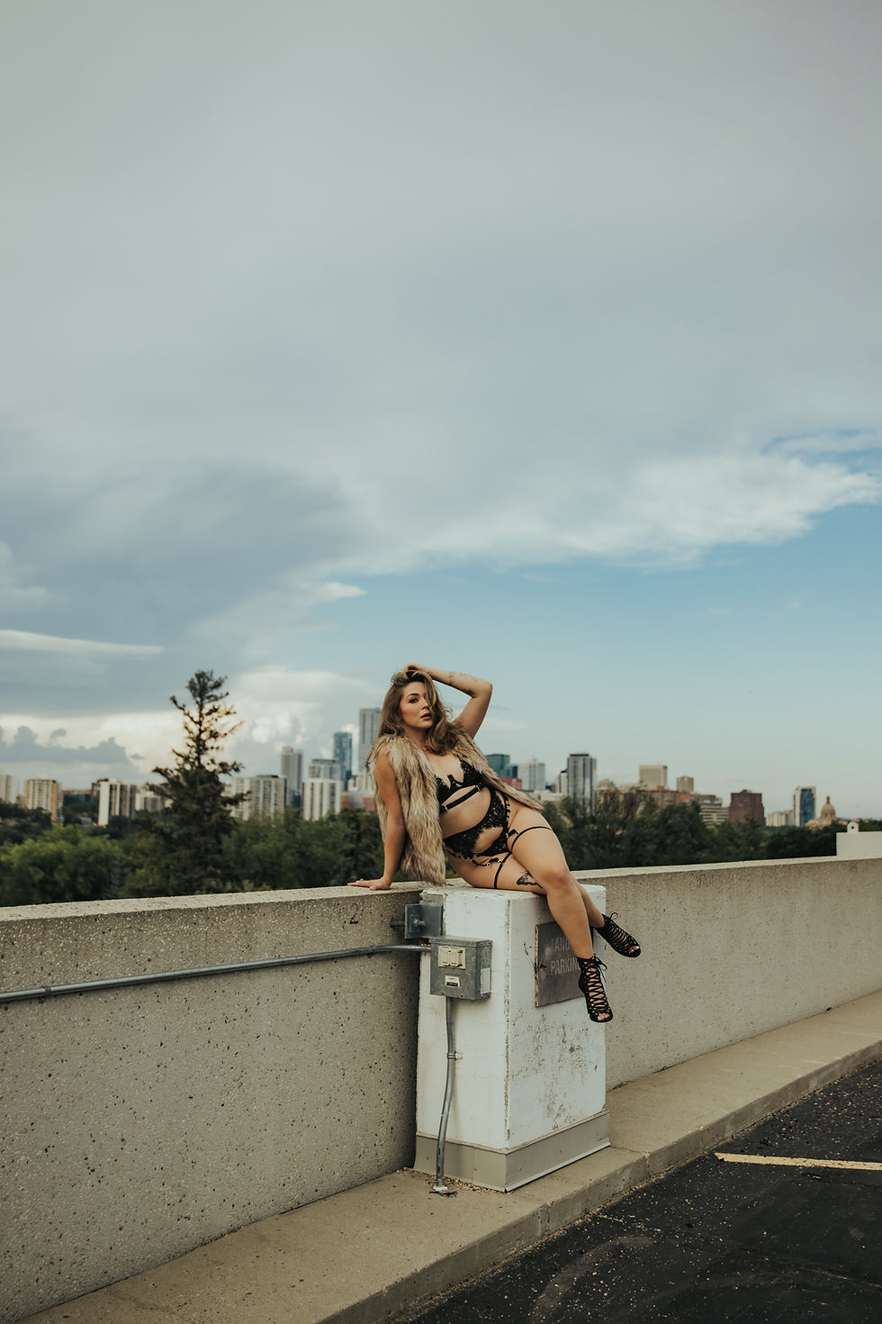 Woman in black lingerie and fur vest poses on a rooftop wall above a city skyline under a cloudy sky.