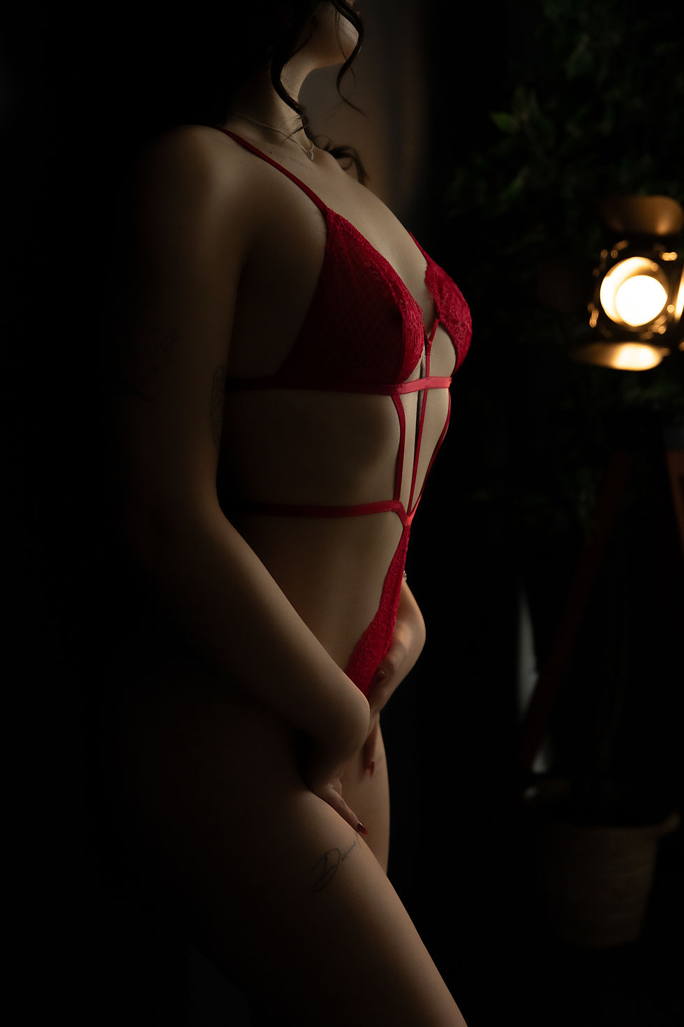Person in red lingerie stands in dim lighting, casting shadows. A soft, warm light in the background creates a moody atmosphere.