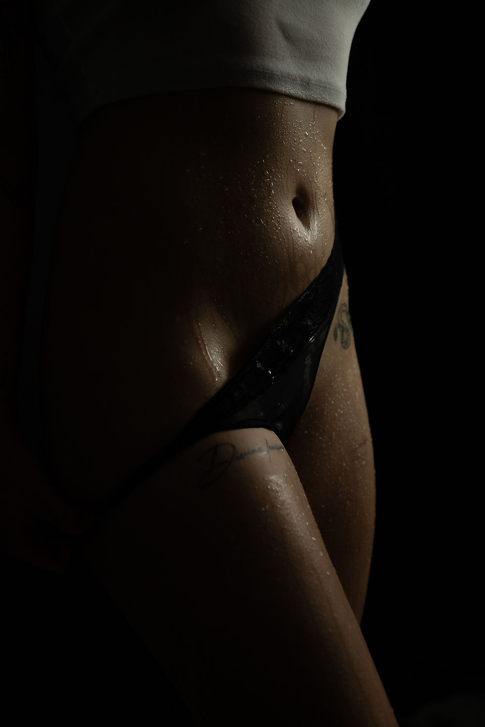 Close-up of a person's midsection in dim light, showing a white top, black underwear, and tattoos with water droplets on the skin.