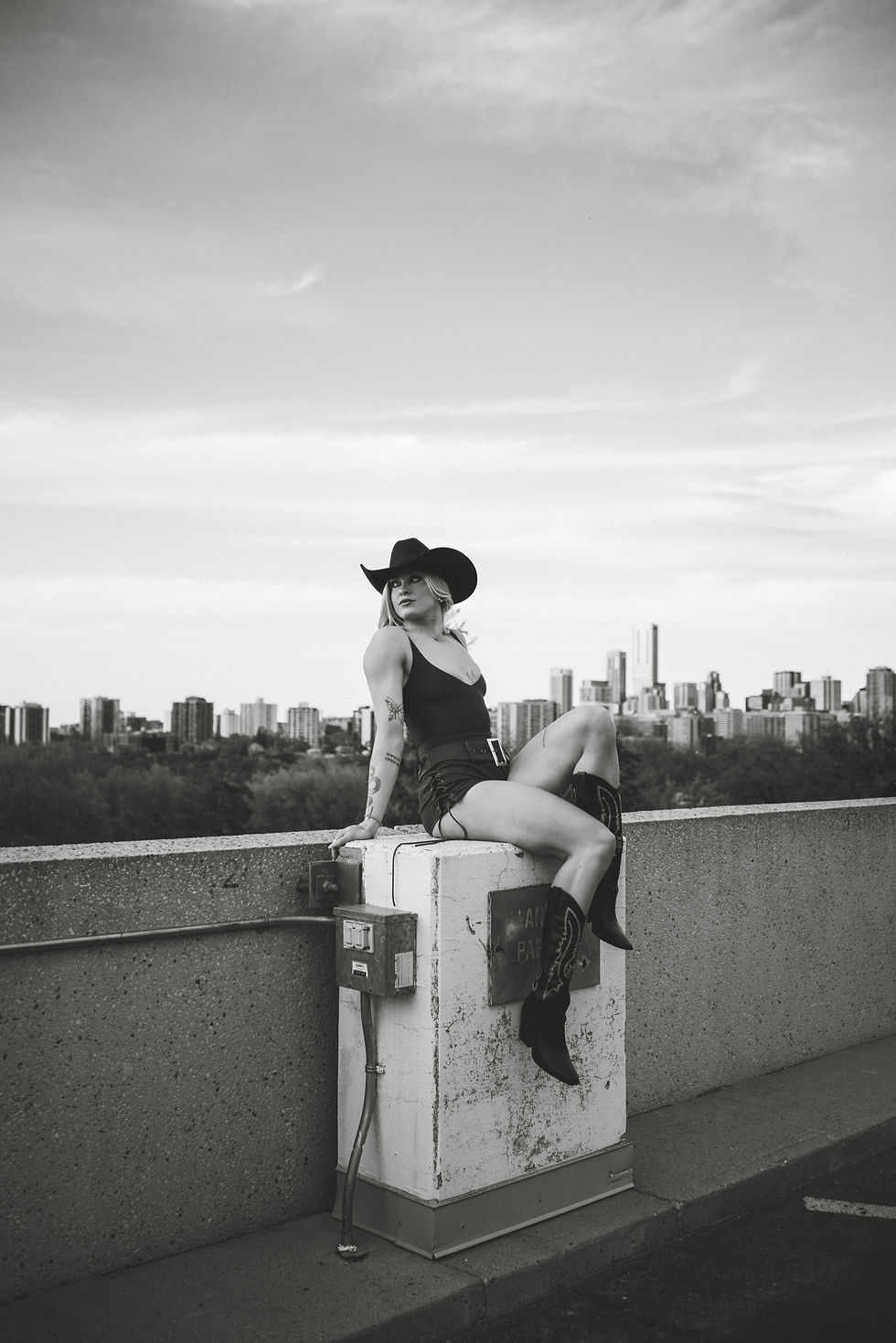 Black-and-white photo of a woman in a cowboy hat and boots sitting on a rooftop ledge, with a city skyline behind her.