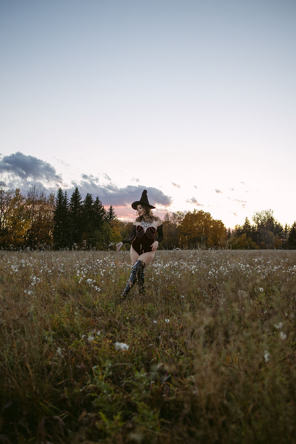 Woman in a black witch hat and costume walks through a wildflower field at sunset, with dark trees and glowing sky behind her.