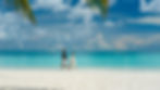 Couple walking hand in hand on a tropical beach with turquoise water, a sailboat in the distance, and palm leaves above. Calm and serene.