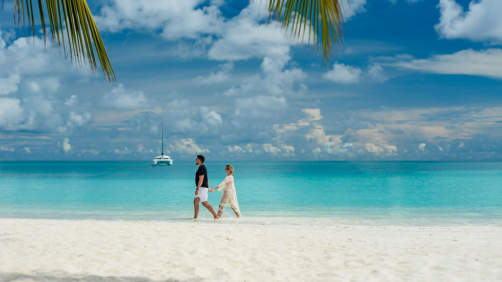 Couple walking hand in hand on a tropical beach with turquoise water, a sailboat in the distance, and palm leaves above. Calm and serene.