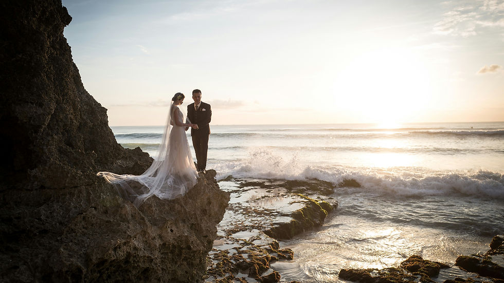 Bride and groom stand on rocky shore, as waves crash around them under a setting sun. The scene is serene and romantic.