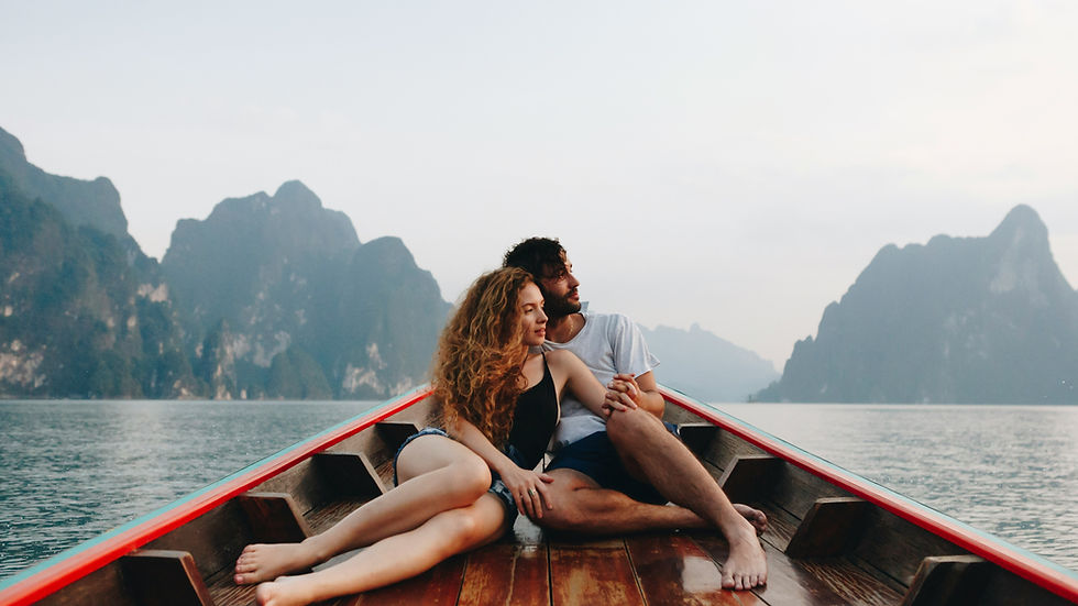 Couple sitting on a wooden boat, holding hands and gazing at serene mountains across a lake. Calm mood with a soft, cloudy sky.