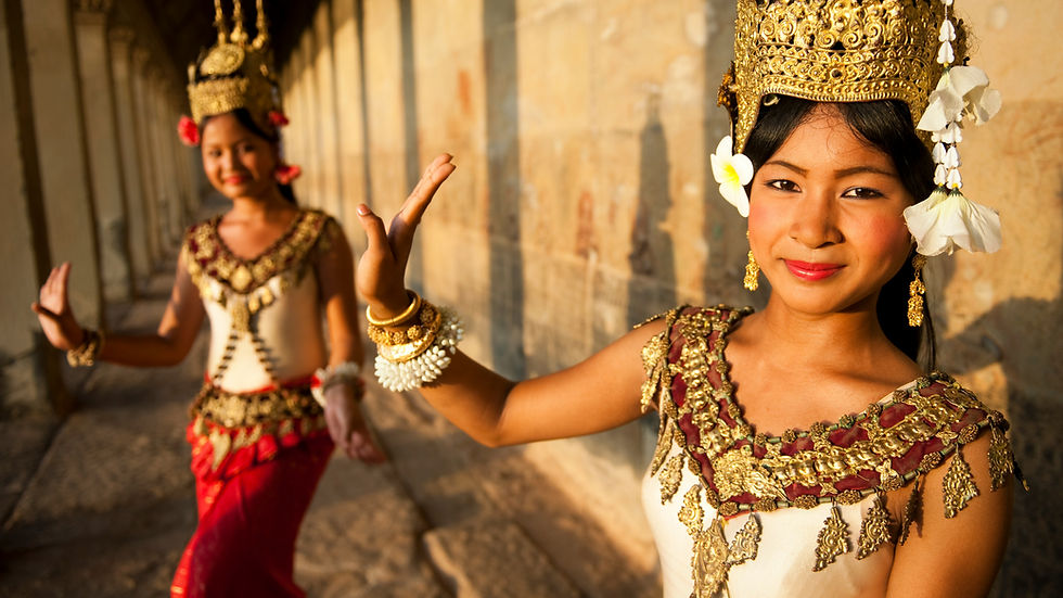 Two women perform a traditional dance in ornate gold costumes, smiling in sunlight against a stone wall background. One is in focus.