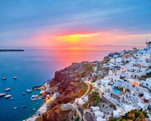 Sunset over Santorini, Greece, with white buildings cascading down cliffs to a calm sea dotted with boats. Vibrant sky in orange and purple hues.
