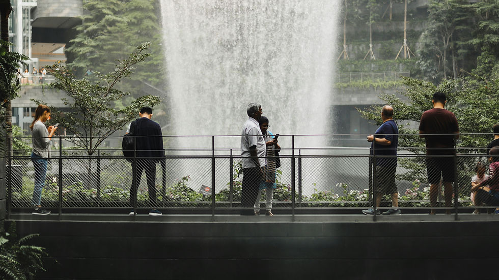 People stand on a viewing platform with smartphones near a large waterfall indoors, surrounded by lush greenery. The mood is calm.
