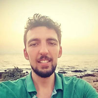 Selfie of a man with short curly hair and a trimmed beard, wearing a green shirt, standing outdoors near a rocky shoreline at