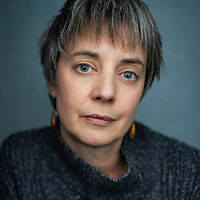 Portrait of a woman with short salt-and-pepper hair and blue eyes, wearing a dark knit sweater and amber earrings, looking ca