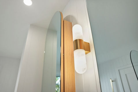 Curved mirror and brushed brass wall light above a custom walnut vanity in a modern coastal bathroom