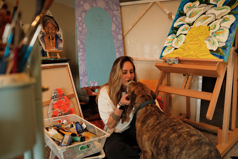 Woman kisses a brindle dog in a cozy art studio, surrounded by easels, paint supplies, and colorful portraits.