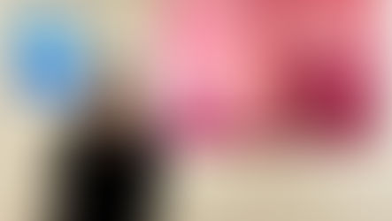 Yi-Chiao Chen in a black coat poses in front of two paintings, one pink and one blue, in a gallery. She smiles, thumb up, against a white wall.