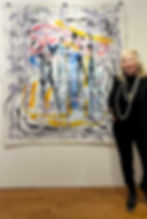 Suzanne in black with pearl necklace stands beside colorful abstract painting on white wall, featuring splashes of purple, yellow, and blue.