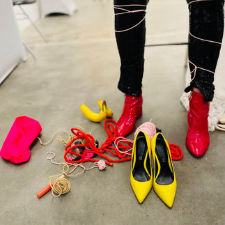A pair of legs wrapped in rope stands beside red and yellow high heels and scattered objects.