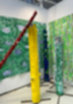 Colorful art installation with painted tree trunks in red, yellow, and blue against a textured green patterned backdrop. Bright and vibrant.