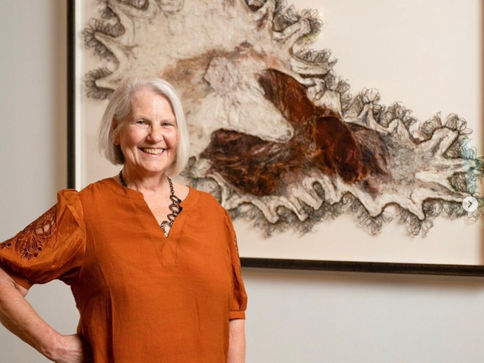 Carla J Fisher in orange blouse stands in front of abstract art with earthy tones and jagged shapes. White wall background.