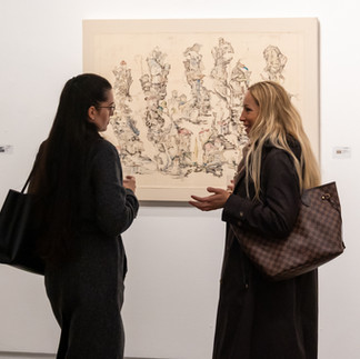 Two women standing in front of a large, textured drawing by Denny Theocharakis, discussing the artwork during the exhibition