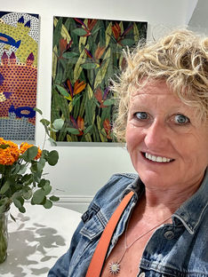 Smiling Julia in denim jacket beside colorful art and orange flowers. Bright, cheerful gallery setting with patterned and floral paintings.