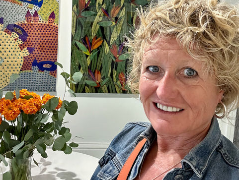 Smiling Julia in denim jacket beside colorful art and orange flowers. Bright, cheerful gallery setting with patterned and floral paintings.