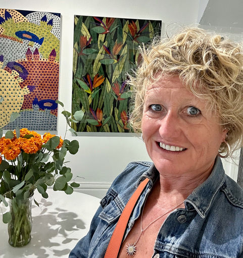 Smiling Julia in denim jacket beside colorful art and orange flowers. Bright, cheerful gallery setting with patterned and floral paintings.