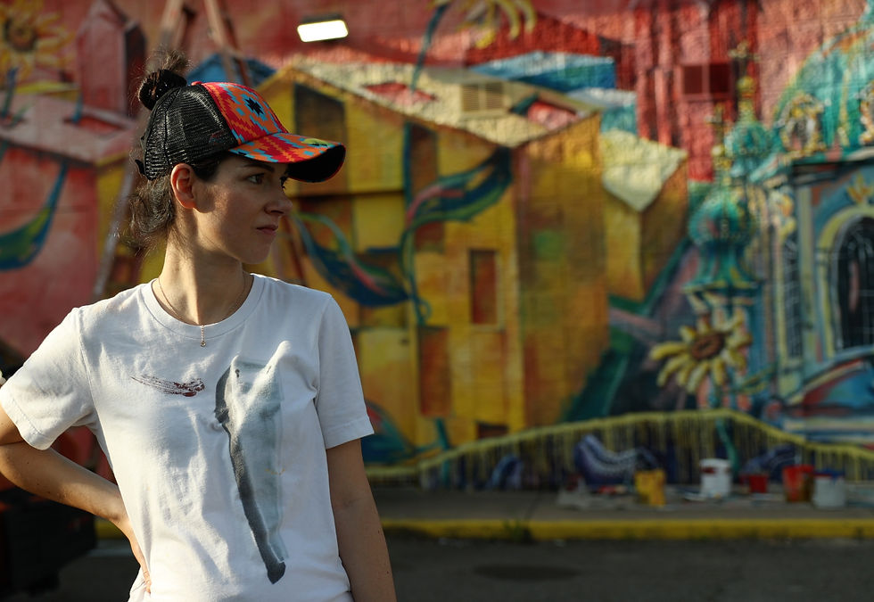 Raisa Nosova in colorful cap and white shirt stands in front of a vibrant mural. Sunlight casts shadows. Buckets and painting tools on ground.
