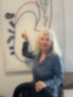 Suzanne with long hair, pointing at colorful bead artwork on the wall. She wears a patterned blouse and smiles warmly in a well-lit room.