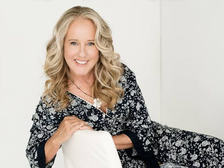 Jean Nestares smiling while seated against a light neutral background, wearing a black floral dress and leaning gently on a white chair.