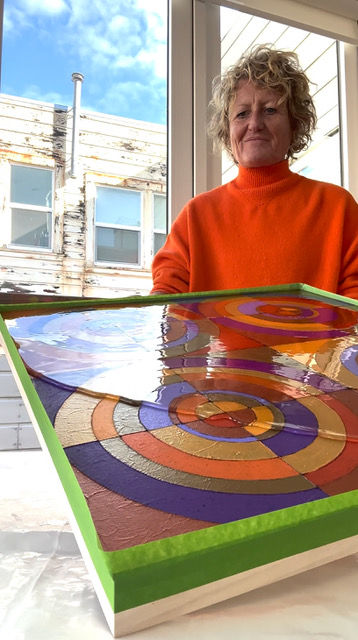 Julia in an orange sweater stands by a colorful painting with concentric circles, framed in green. Sunlit room with windows in the background.