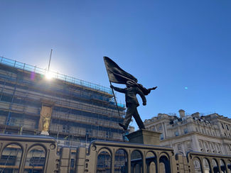 A statue of a man with a waving cape stands on a pedestal against a clear blue sky. Scaffolding and historical buildings are in the background.