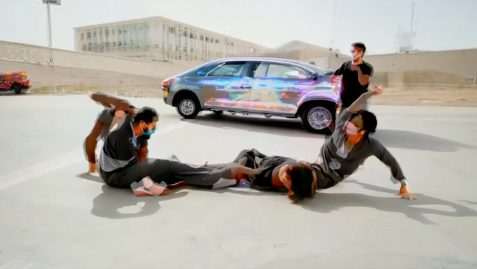People in masks dance on an urban street, with a car nearby. Art exhibition at NEW MUSEUM