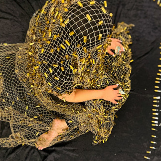 An artist covered in gold fabric with bullets sits curled up on a black background.
