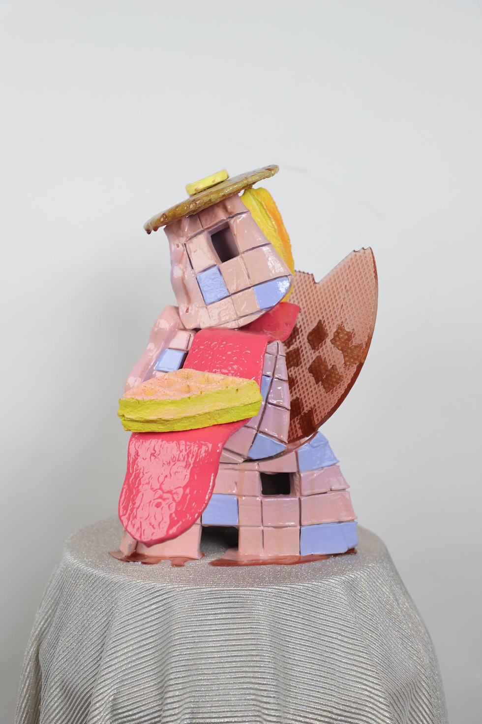 Colorful abstract sculpture of interlocking shapes and blocks, with pink, yellow, and red tones, set on a textured gray base.