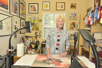 Smiling Bev Yankwitt works on an abstract painting in a bright art studio, surrounded by framed art, brushes, and award ribbons.