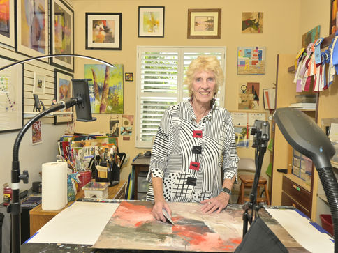 Smiling Bev Yankwitt works on an abstract painting in a bright art studio, surrounded by framed art, brushes, and award ribbons.