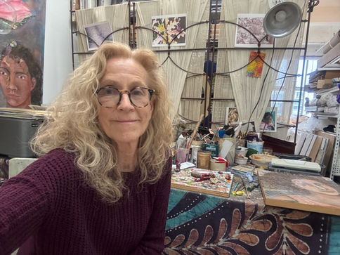 Portrait of an artist seated in her studio, surrounded by painting tools, collage materials, artworks, shelves, and a worktable filled with creative supplies.