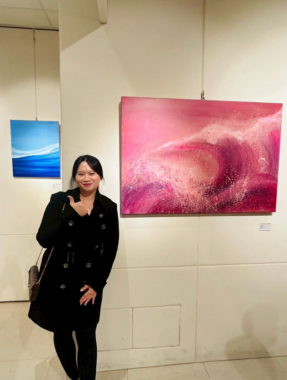 Yi-Chiao Chen in a black coat poses in front of two paintings, one pink and one blue, in a gallery. She smiles, thumb up, against a white wall.