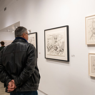 A man with his hands behind his back looks closely at framed factory-themed drawings by Denny Theocharakis displayed on a white gallery wall