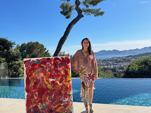 Eva March in patterned dress stands by a pool holding a vivid red abstract painting, with sea and hills behind.