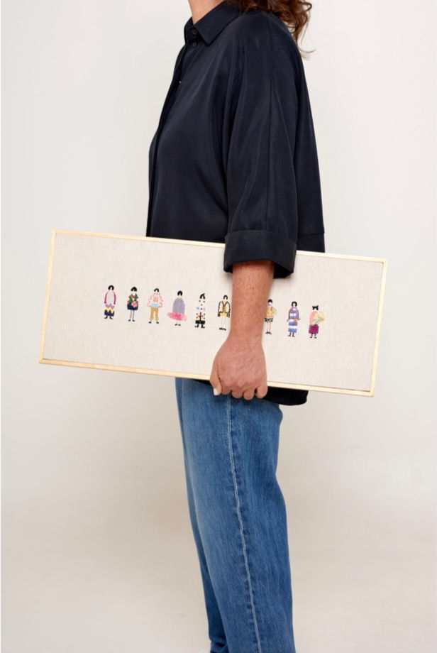 Andrea Bruder in a dark shirt and jeans holds a framed artwork featuring small, colorful figures. The background is plain and light-colored.