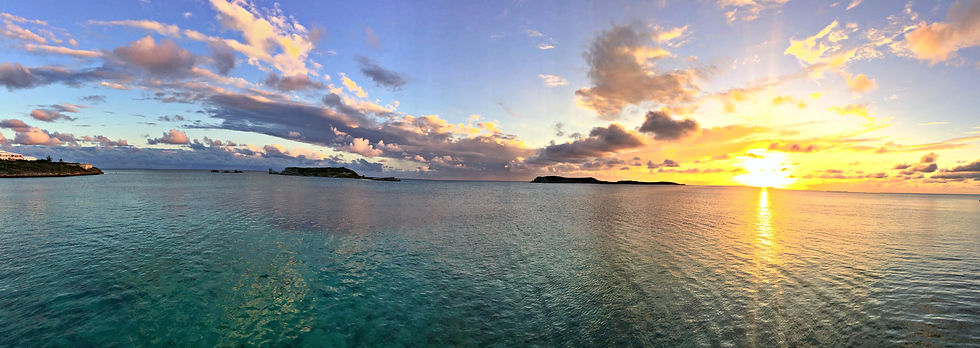 Sunset over a few small, rocky islands in the ocean. Clouds are cast gold, pink, and purple, water is dark turquoise.