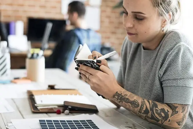 Woman with tattoos writes in a notebook at a desk with papers and a phone. Background shows a blurred person and a brick wall.