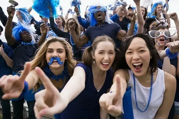 A group of enthusiastic fans in blue apparel cheer, some wearing wigs and face paint, with festive expressions in a crowded stadium setting.