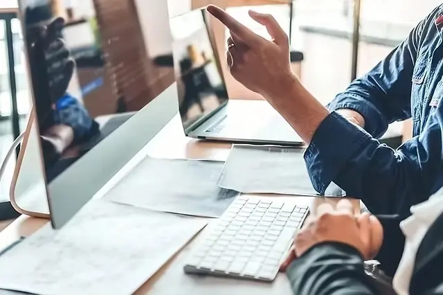 Person pointing at computer screen with code, seated at desk with keyboard and papers. Casual setting, focused mood.