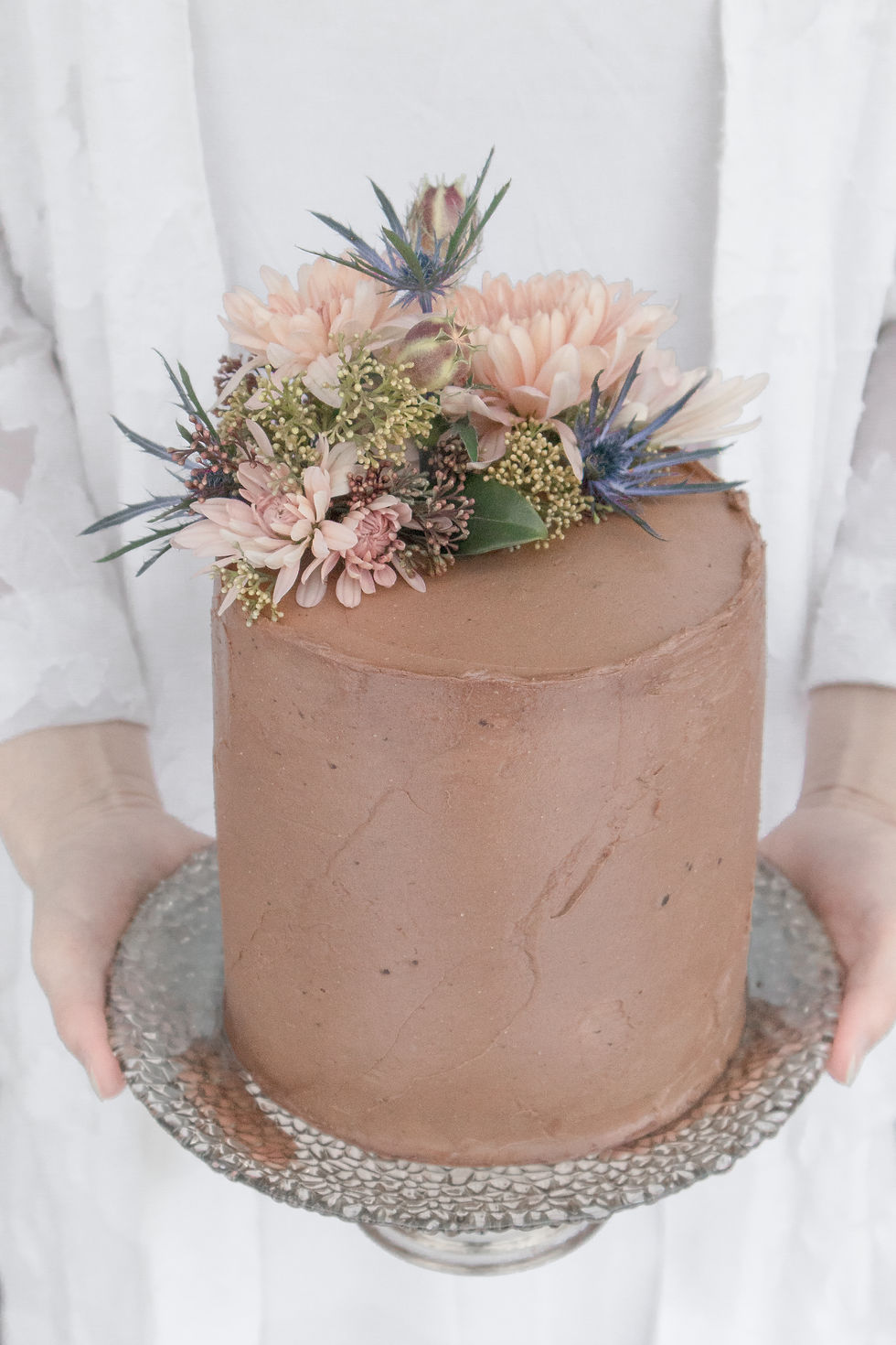 Top 10 cakes. Top 10 cake recipes. Beautiful food photography of a Roasted Hazelnut, Dark Chocolate & Raspberry Cake. Flat lay with pink flowers and coffee beans. Young woman holding cake decorated with modern flowers, wearing a white slip dress and kimono, both from Aritzia.