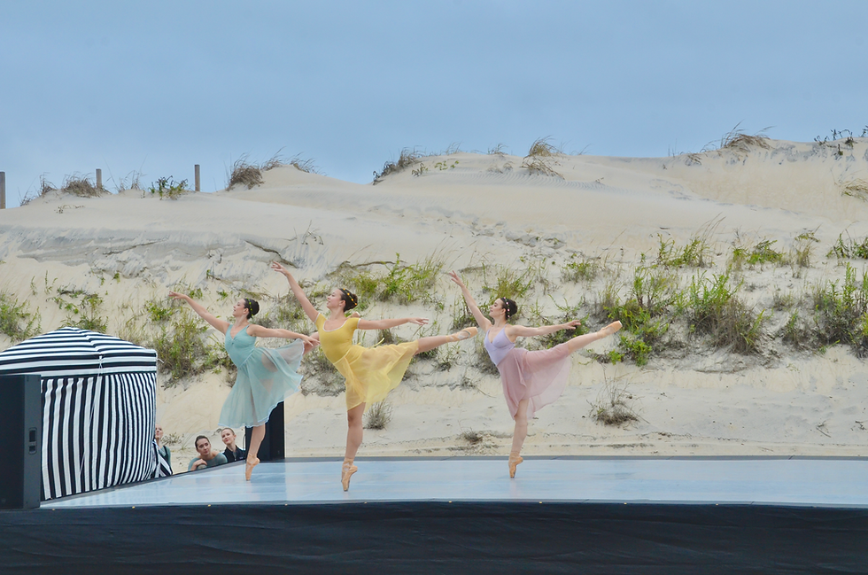 Three ballerinas in pastel tutus perform arabesques on an outdoor stage before sandy dunes and a clear blue sky.