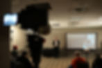 Speaker presenting in a dimly lit room to a seated audience. A camera in the foreground; screen displays "2 Things Everyone Needs." Education and breakout sessions for growth at Wash and Learn Midwest. Pressure washing and soft washing education event in Chicago.